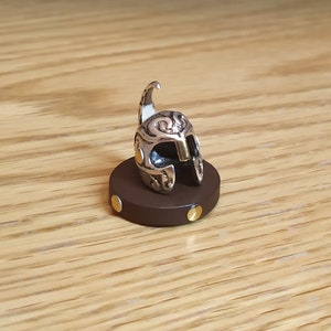 May include: A silver and gold miniature helmet with intricate details sits on a brown circular base with gold accents.
