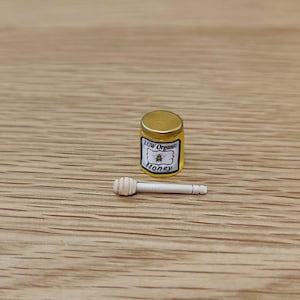 May include: A small glass jar of honey with a yellow label that reads "LGW Organic Honey" and a small illustration of a bee. A wooden honey dipper is next to the jar.