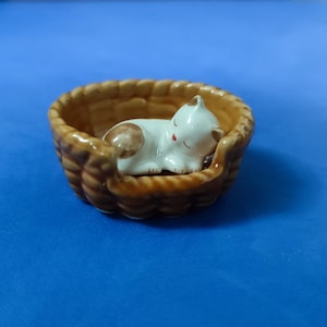 May include: A white and brown ceramic cat figurine sleeping in a brown wicker basket.