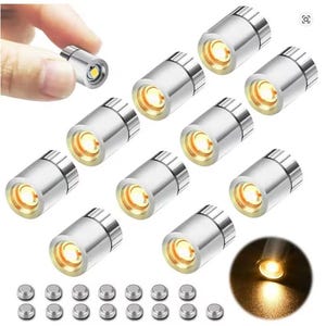 May include: A collection of small, silver-colored LED lights with a warm yellow glow. The image shows a hand holding one light, with several others arranged in a group. Small, silver batteries are also visible.