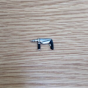 May include: A silver-colored drill with a black handle. The drill bit is visible at the end of the tool. The drill is set against a light brown wooden background.