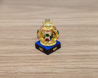 May include: A miniature gold crown with red, green, and white gemstones sits on a blue velvet base. The crown is a decorative piece and is likely made of metal.
