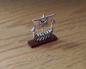 May include: A silver miniature Viking ship with a wooden base. The ship has a sail with a stylized animal design.
