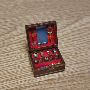 May include: A small wooden jewellery box with a red velvet interior. The box is open and contains a variety of jewellery, including earrings, necklaces, and rings. There is also a small mirror and a pair of scissors inside the box.