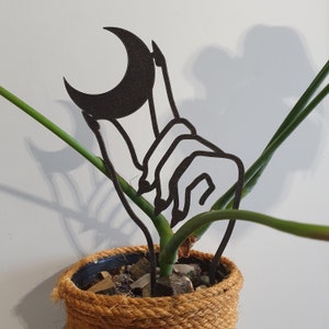 May include: Black metal plant stake shaped like a hand holding a crescent moon. The stake is inserted into a pot with a green plant and brown rope around the rim.