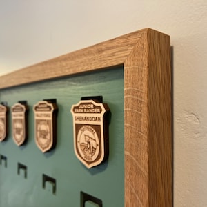 May include: A wooden frame with a green background and four wooden badges. The badges are for Junior Park Rangers at Shenandoah National Park. The frame is made of light brown wood.