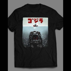 Può includere: Maglietta nera con una grafica di Godzilla che emerge dall'acqua con una barca sopra. Il testo "ゴジラ" è scritto in rosso sopra l'immagine.
