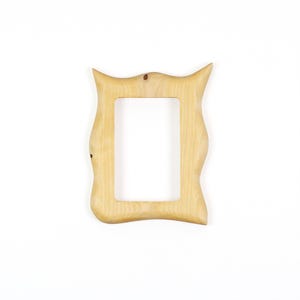Pu&ograve; includere: Una cornice portafoto in legno di colore chiaro con venature naturali. La cornice presenta un bordo esterno irregolare e ondulato con due sporgenze appuntite agli angoli superiori e un'apertura centrale rettangolare per una foto.
