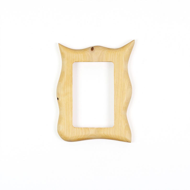 Pu&ograve; includere: Una cornice portafoto in legno di colore chiaro con venature naturali. La cornice presenta un bordo esterno irregolare e ondulato con due sporgenze appuntite agli angoli superiori e un'apertura centrale rettangolare per una foto.