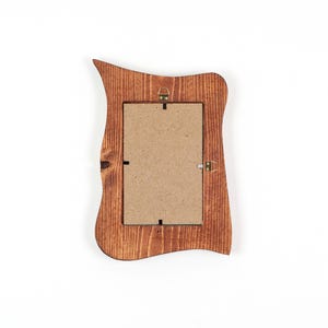 Peut inclure: Un cadre photo rectangulaire en bois avec un bord ext&eacute;rieur irr&eacute;gulier et ondul&eacute;, dot&eacute; d'une finition teint&eacute;e brun fonc&eacute;. L'arri&egrave;re du cadre pr&eacute;sente un panneau de fibres brun clair fix&eacute; par des clips m&eacute;talliques et une attache murale en laiton.