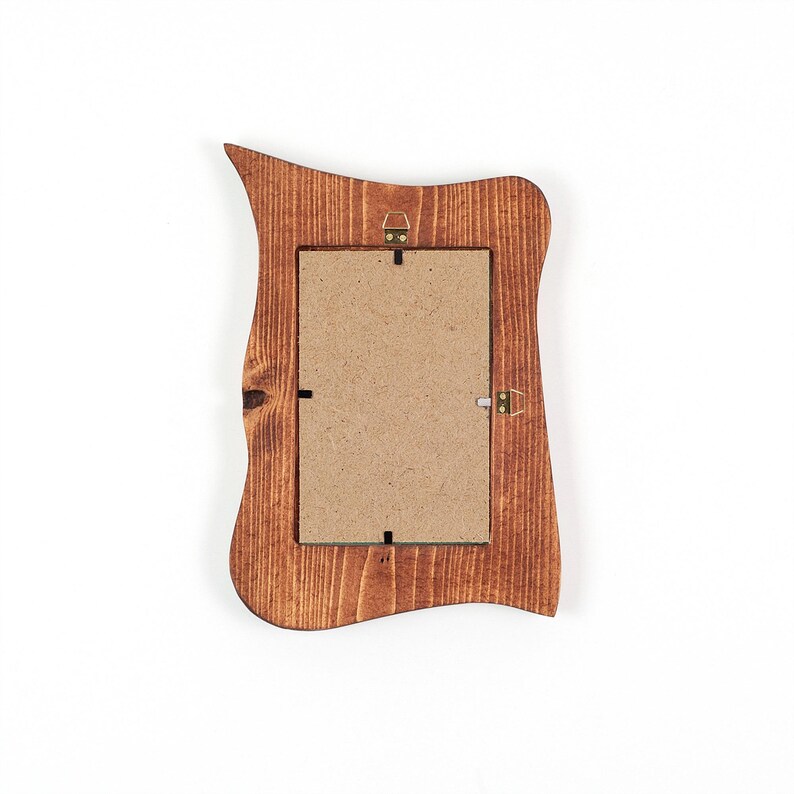 Peut inclure: Un cadre photo rectangulaire en bois avec un bord ext&eacute;rieur irr&eacute;gulier et ondul&eacute;, dot&eacute; d'une finition teint&eacute;e brun fonc&eacute;. L'arri&egrave;re du cadre pr&eacute;sente un panneau de fibres brun clair fix&eacute; par des clips m&eacute;talliques et une attache murale en laiton.
