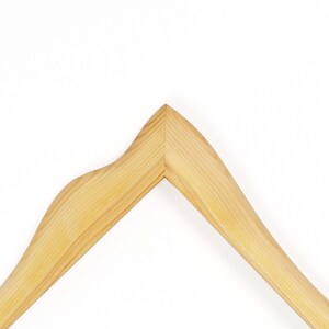 May include: A light-coloured wooden clothes hanger featuring a smooth, natural grain finish. The hanger has a distinct, angular peak at the centre where the two arms meet, with a curved shoulder contour on the left side and a straight slope on the right.