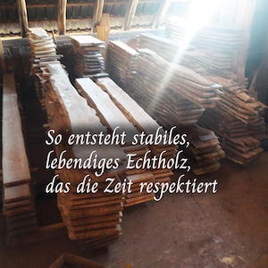 K&ouml;nnte beinhalten: Mehrere Stapel aus rohen, unbehandelten Holzbohlen mit nat&uuml;rlichen Baumkanten in einer Werkstatt. Der Text lautet: So entsteht stabiles, lebendiges Echtholz, das die Zeit respektiert. Das Holz zeigt verschiedene Braunt&ouml;ne und Maserungen.