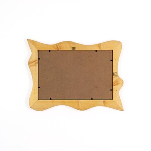 May include: Back view of a light-colored wooden picture frame with a wavy, irregular edge. The frame has a brown backing and small metal hangers. The frame is designed to hold a rectangular photo or artwork.