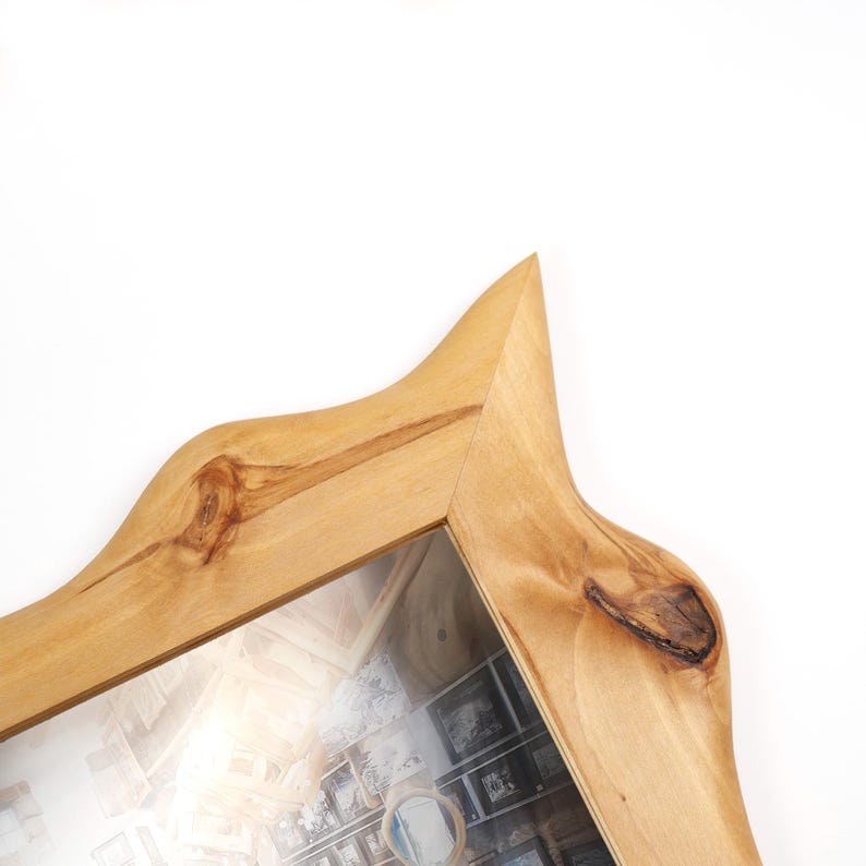 May include: A light brown wooden mirror frame with a natural wood grain finish and visible knots. The frame features an irregular, pointed top edge and a smooth, polished surface, showcasing the organic texture and warm tones of the wood.