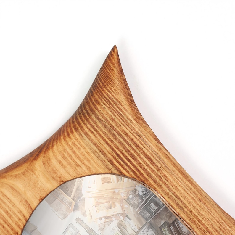 May include: A wooden mirror with a distinctive, curved frame. The frame is crafted from light brown wood, showcasing its grain, and the mirror has a diamond-like shape. The mirror reflects a room with various objects.