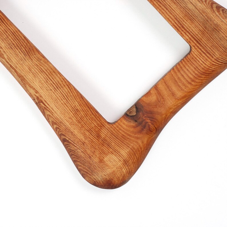 May include: Close-up of a wooden frame with a square aperture. The frame is made of light brown wood with visible grain and a smooth, curved corner. The wood has a natural, warm tone.