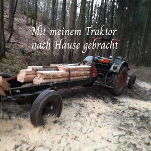 May include: A blue tractor pulling a black trailer loaded with stacked light-coloured wooden planks in a forest setting. The text "Mit meinem Traktor nach Hause gebracht" is overlaid in white script across the upper portion of the image.