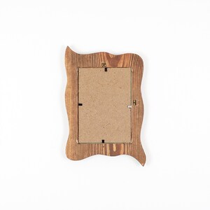 Peut inclure: Un cadre photo rectangulaire en bois avec un bord ondul&eacute; et organique. Le cadre pr&eacute;sente un motif de grain de bois brun visible et un dos en panneau de fibres brun clair fix&eacute; par de petites attaches m&eacute;talliques et une attache de suspension.