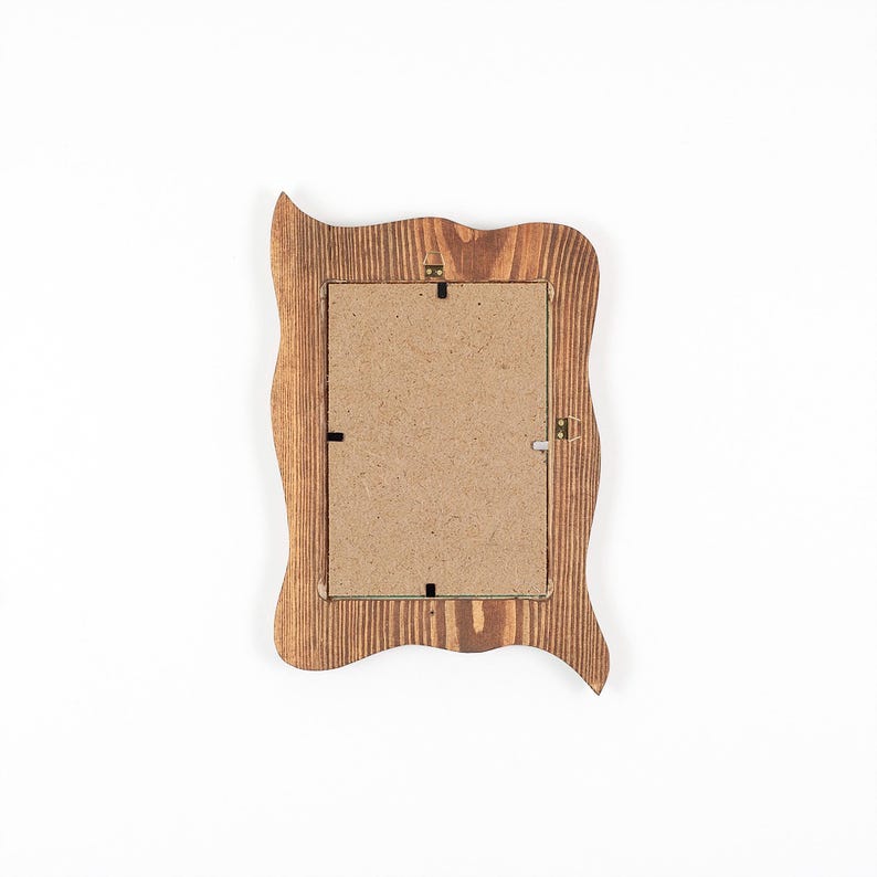 Peut inclure: Un cadre photo rectangulaire en bois avec un bord ondul&eacute; et organique. Le cadre pr&eacute;sente un motif de grain de bois brun visible et un dos en panneau de fibres brun clair fix&eacute; par de petites attaches m&eacute;talliques et une attache de suspension.