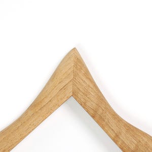 Puede incluir: Una percha de madera con acabado natural. La percha tiene un dise&ntilde;o curvo y una parte superior triangular, con un color marr&oacute;n claro y liso. La percha est&aacute; sobre un fondo blanco.
