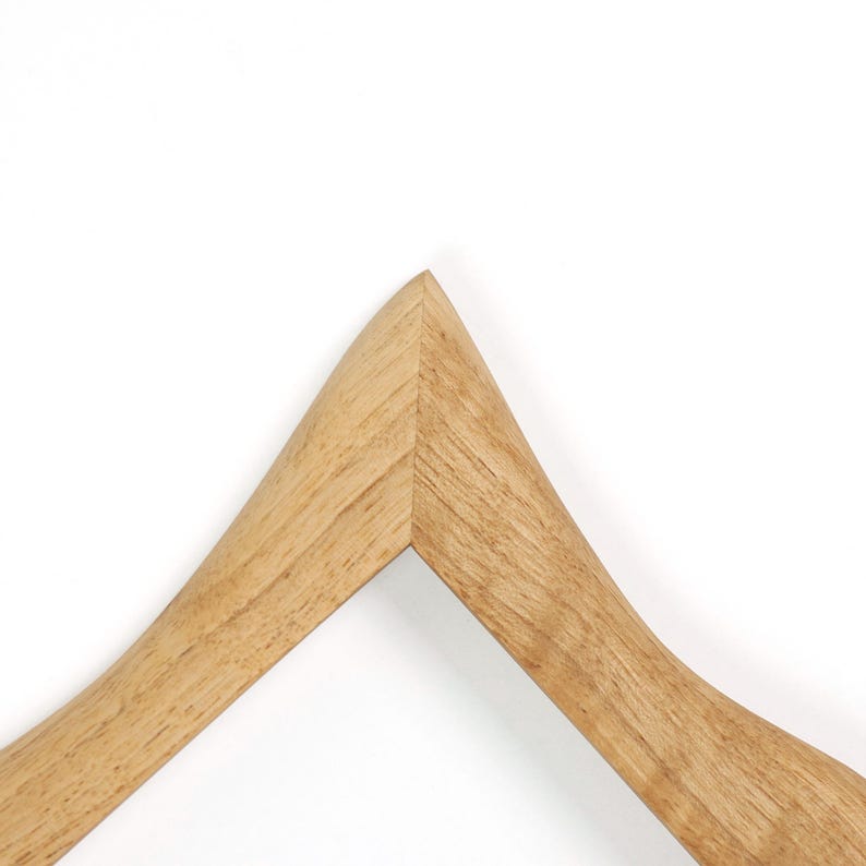 Puede incluir: Una percha de madera con acabado natural. La percha tiene un dise&ntilde;o curvo y una parte superior triangular, con un color marr&oacute;n claro y liso. La percha est&aacute; sobre un fondo blanco.