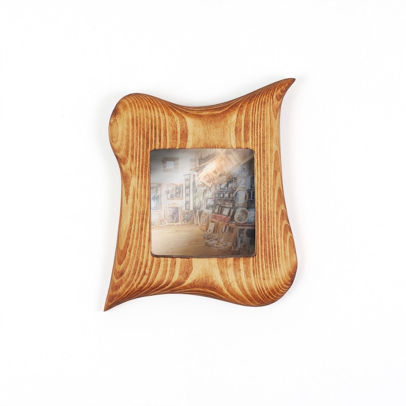 Puede incluir: Un marco de fotos cuadrado de madera con un acabado de veta de madera marr&oacute;n claro natural y un borde exterior asim&eacute;trico y ondulado. El marco presenta una abertura cuadrada central que muestra una peque&ntilde;a imagen de un interior de tienda.