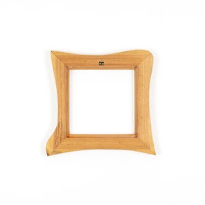 Puede incluir: Un marco de fotos cuadrado de madera con un acabado marr&oacute;n claro. El marco tiene un dise&ntilde;o curvo &uacute;nico en los bordes exteriores. Un peque&ntilde;o colgador de metal est&aacute; fijado en la parte posterior. El interior del marco es blanco.