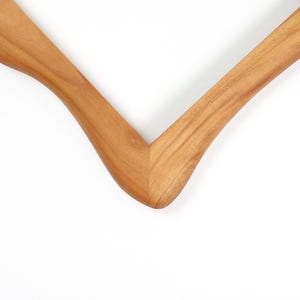 Op de afbeelding: Close-up van een houten kledinghanger. De hanger is gemaakt van lichtbruin hout en heeft een glad, gebogen ontwerp. De hanger staat tegen een witte achtergrond.