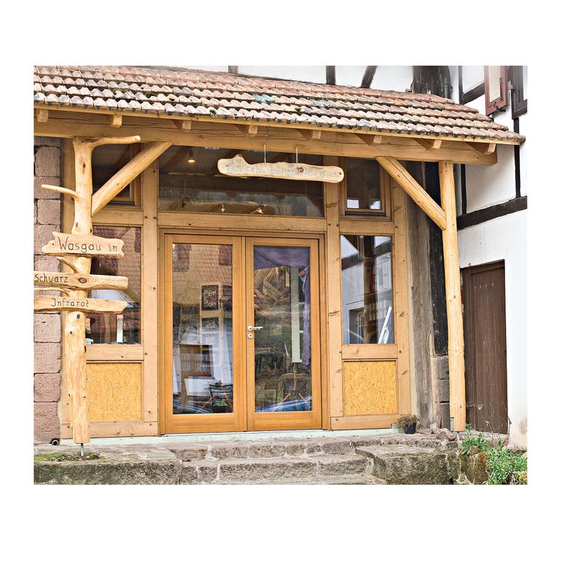May include: Exterior view of a building with a wooden frame and a tiled roof. The entrance features double glass doors and a sign that reads "Wasgau in Schwarz Infrared". A wooden signpost points towards different directions.