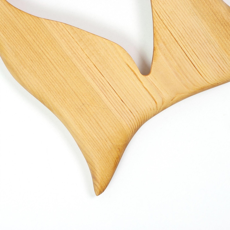 Op de afbeelding: Een lichtgekleurd houten object met een gladde, gebogen en puntige vorm. Het oppervlak vertoont een natuurlijk nerfpatroon met zichtbare lijnen die langs de lengte van het hout lopen, inclusief een duidelijke V-vormige uitsparing bovenaan.