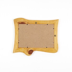 Puede incluir: Un marco de fotos rectangular hecho de madera natural de color claro con un acabado de borde vivo. El marco presenta un nudo de madera oscura prominente en el borde inferior, un respaldo de tablero de fibra color canela y clips met&aacute;licos.