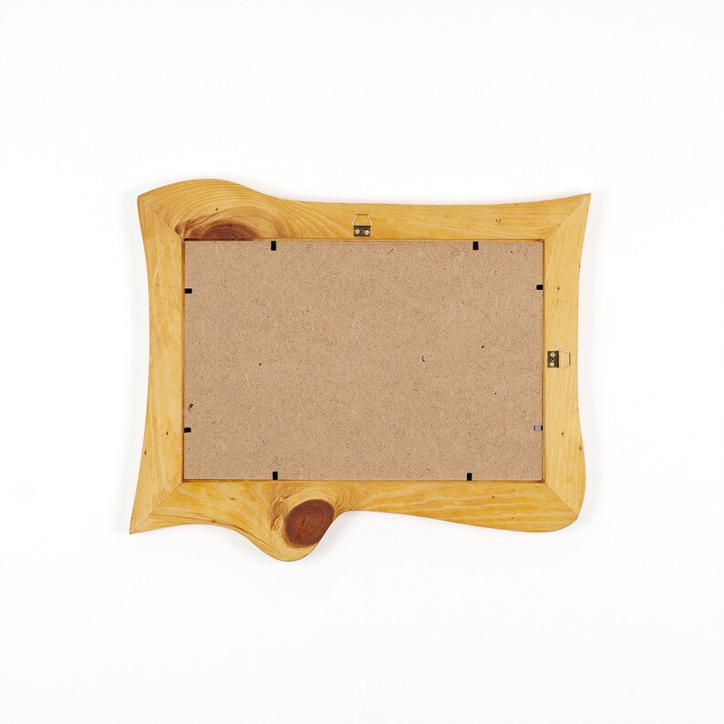 Puede incluir: Un marco de fotos rectangular hecho de madera natural de color claro con un acabado de borde vivo. El marco presenta un nudo de madera oscura prominente en el borde inferior, un respaldo de tablero de fibra color canela y clips met&aacute;licos.