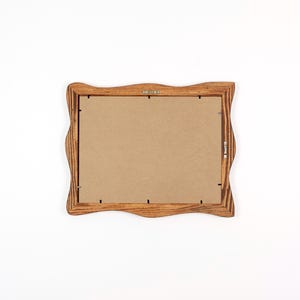 May include: A wooden picture frame with a wavy edge design. The frame is made of light brown wood and has a cardboard backing. The frame has metal hardware for hanging.