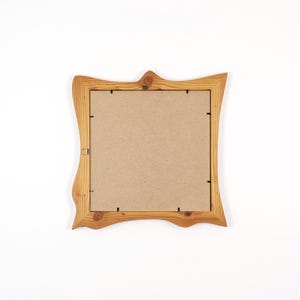 May include: A square, light brown wooden picture frame with a decorative, wavy edge. The frame has a natural wood grain and a cardboard backing. A small metal hanger is attached to the top center.