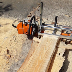 Puede incluir: Un accesorio de aserradero port&aacute;til para motosierra con un marco de metal plateado y rieles gu&iacute;a verticales, montado sobre una motosierra naranja y blanca, posicionado sobre una tabla de madera clara con veta y textura visibles.