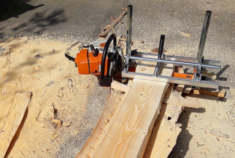 Puede incluir: Un accesorio de aserradero port&aacute;til para motosierra con un marco de metal plateado y rieles gu&iacute;a verticales, montado sobre una motosierra naranja y blanca, posicionado sobre una tabla de madera clara con veta y textura visibles.
