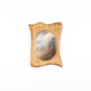 Op de afbeelding: Een lichtbruine houten fotolijst met een uniek, golvend ontwerp. De lijst heeft een ovale opening die een kleurrijke afbeelding van een kamer vol kunst toont. De lijst staat tegen een witte achtergrond.