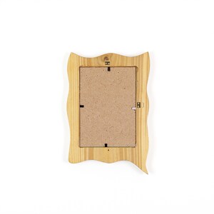 May include: A light-colored wooden picture frame with a wavy, decorative edge. The back of the frame is visible, showing a cardboard backing and metal hardware for hanging. The frame is designed to hold a rectangular photo.