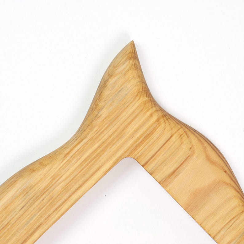 Pu&ograve; includere: Primo piano di un'appendiabiti in legno con finitura a venatura naturale. L'appendiabiti ha un design curvo con una parte superiore a punta e un ritaglio per il collo di un capo. Il legno &egrave; marrone chiaro.