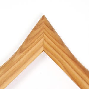 May include: Close-up of a wooden picture frame corner. The frame is made of light-colored wood with visible grain. The corner is a sharp, right angle, and the frame has a curved design. The background is plain white.