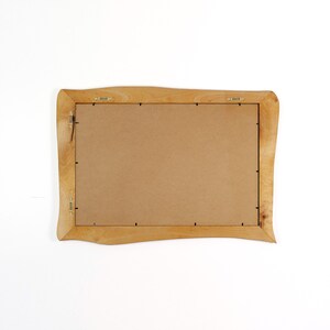 Peut inclure: Un cadre photo rectangulaire en bois avec une finition brun clair. Le cadre a une forme ondul&eacute;e et organique et un dos brun. Le mat&eacute;riel de suspension en m&eacute;tal est fix&eacute; &agrave; l'arri&egrave;re.