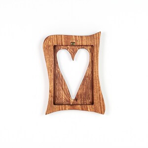 May include: A wooden picture frame with a heart-shaped cutout. The frame is a warm brown color and has a wavy, decorative edge. A small metal hanger is attached to the top for wall mounting.