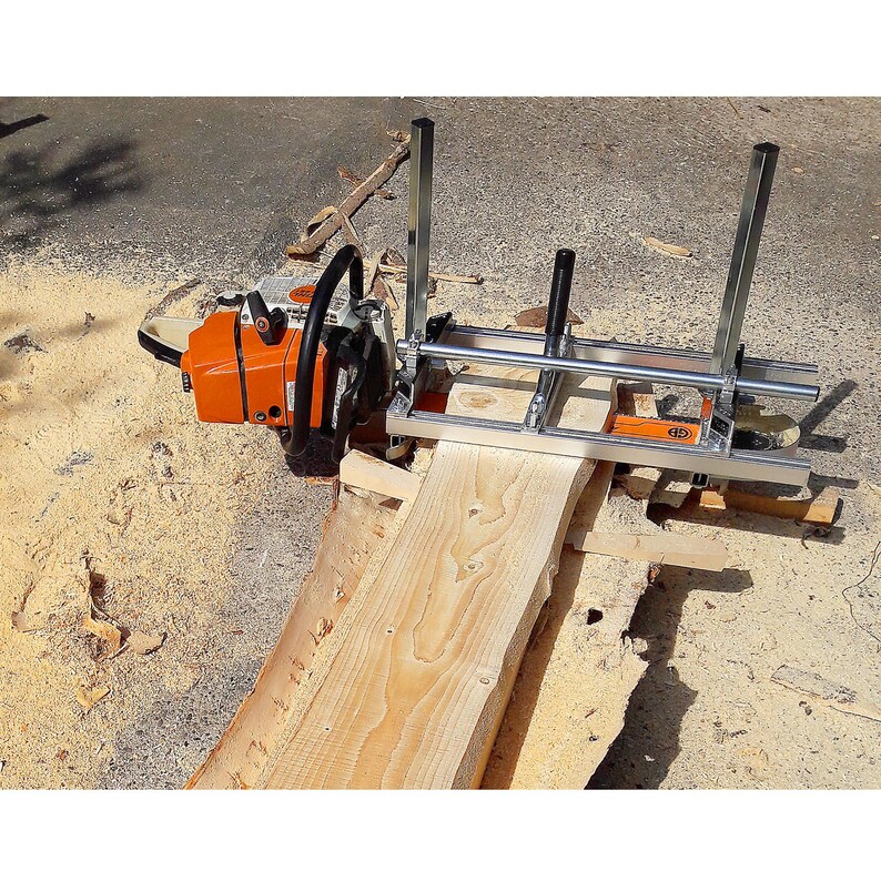 Peut inclure: Une tron&ccedil;onneuse orange et noire fix&eacute;e &agrave; un guide de fraisage en m&eacute;tal, coupant une planche de bois brun clair. L'ensemble est pos&eacute; sur une surface en b&eacute;ton, avec des copeaux de bois &eacute;parpill&eacute;s. La tron&ccedil;onneuse est positionn&eacute;e pour une coupe droite.