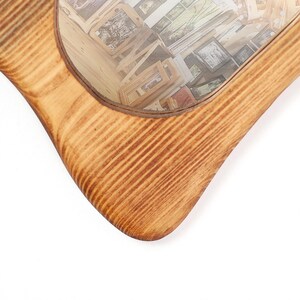May include: A wooden mirror with a light brown finish. The frame has a distinctive, curved shape. The mirror reflects a collection of framed pictures, creating a layered effect. The wood grain is visible on the frame.