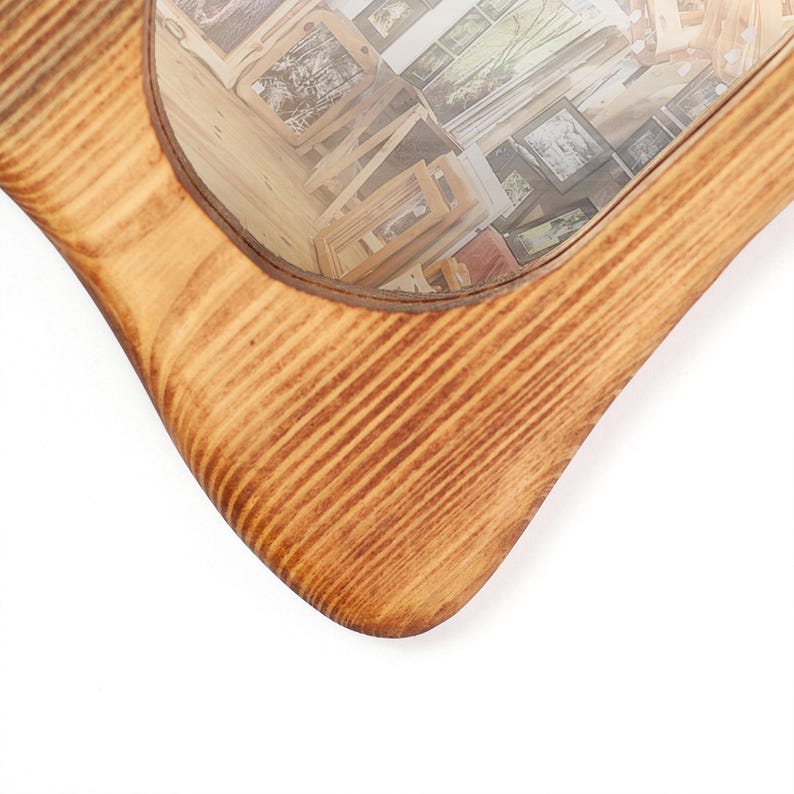 May include: A wooden mirror with a light brown finish. The frame has a distinctive, curved shape. The mirror reflects a collection of framed pictures, creating a layered effect. The wood grain is visible on the frame.