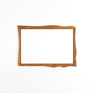 Pu&ograve; includere: Una cornice rettangolare in legno con finitura marrone chiaro. La cornice ha un design a bordo unico e ondulato ed &egrave; vuota, con un interno bianco. L'hardware in metallo &egrave; visibile sul retro per appenderla.