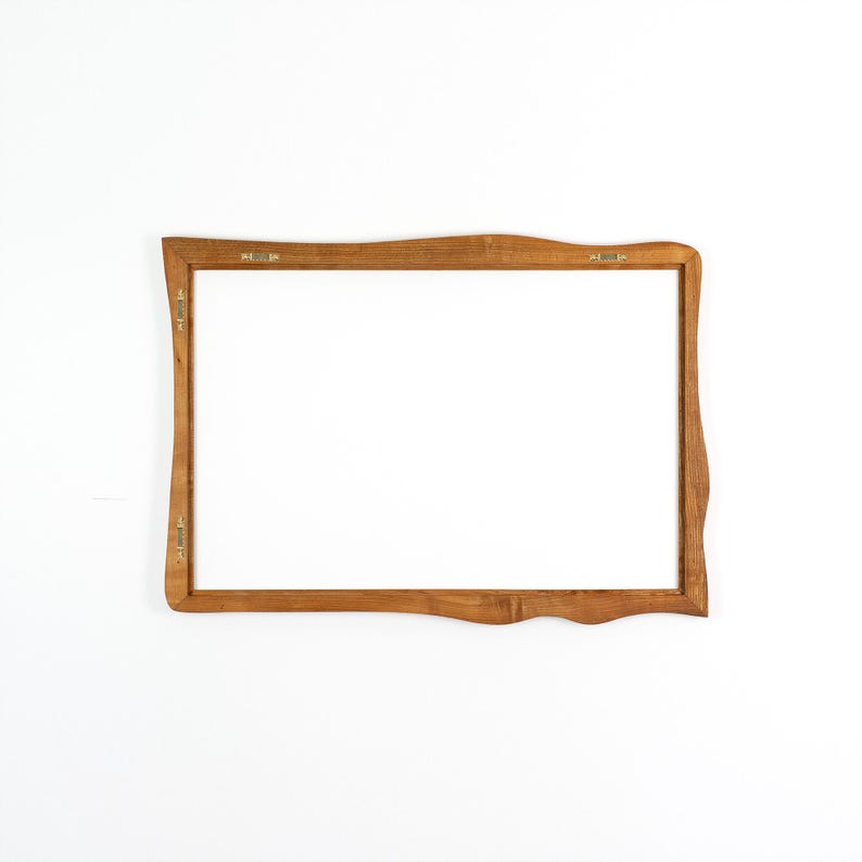 Pu&ograve; includere: Una cornice rettangolare in legno con finitura marrone chiaro. La cornice ha un design a bordo unico e ondulato ed &egrave; vuota, con un interno bianco. L'hardware in metallo &egrave; visibile sul retro per appenderla.