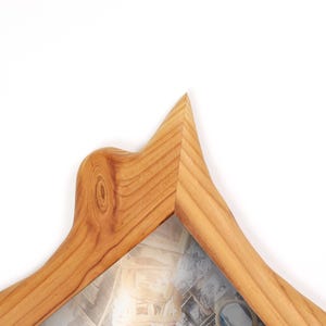 Pu&ograve; includere: Una cornice portafoto in legno marrone chiaro con venature naturali e un bordo superiore appuntito e irregolare. La cornice presenta un nodo visibile nelle venature del legno sul lato sinistro e contiene una fotografia con vari oggetti.
