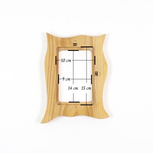 May include: A light-colored wooden picture frame with a wavy, organic outer edge. The frame features internal dimensions marked with black lines and text indicating measurements of 10 cm, 9 cm, 14 cm, and 15 cm. Metal hanging hardware is attached.